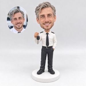 May include: A custom bobblehead figurine of a person with light brown hair, wearing a white button-down shirt, a black tie, and dark trousers. The figure holds a small black car key fob in one hand and stands on a round white base.