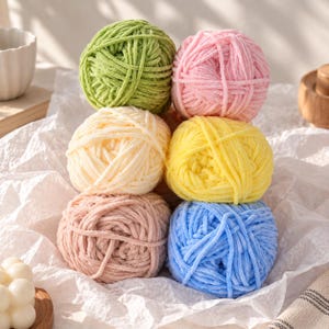 May include: A collection of six round balls of soft, plush yarn arranged in a stack. The colors include light green, pastel pink, cream, bright yellow, dusty rose, and sky blue. Each ball features a thick, velvety texture and a consistent winding.