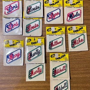 May include: A collection of vintage embroidered name patches on yellow backing cards labeled The Show Offs Emblems. Each rectangular patch features a name in cursive script, including Lori, Louis, Lynn, Marla, Mary, Marty, Matthew, Mike, and Mitchell.