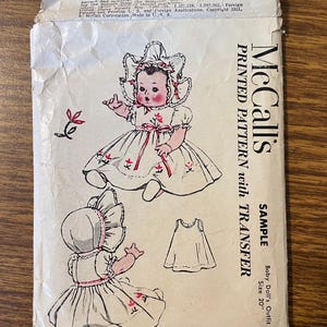 May include: A vintage McCall's printed pattern envelope for a 20 inch baby doll outfit. The paper envelope features black and white illustrations of a doll wearing a dress and bonnet with pink floral accents, along with text including McCall's.