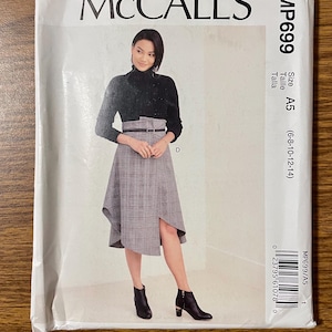 May include: A McCall's sewing pattern envelope, model MP699, size A5 (6-8-10-12-14). The front displays a black long-sleeve turtleneck top paired with a grey plaid high-waisted midi skirt featuring a thin black belt and an asymmetrical hemline.