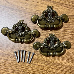 May include: Three antique brass drawer pulls with decorative pineapple and scroll designs. The pulls have curved handles and are accompanied by five silver screws, perfect for furniture restoration or DIY projects.
