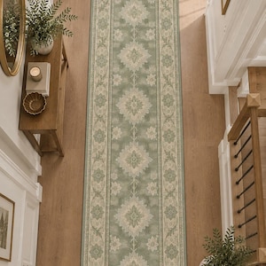May include: A long rectangular runner rug featuring a sage green center field with a repeating cream-colored floral medallion pattern. The rug is framed by a wide cream border decorated with intricate green botanical motifs and a thin outer edge.