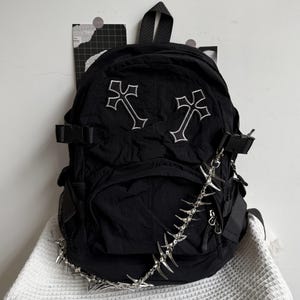 Può includere: Uno zaino nero con due toppe a forma di croce ricamate in bianco sulla tasca anteriore. La borsa include cinghie di compressione laterali con fibbie in plastica nera e una catena decorativa in metallo argentato con punte acuminate.