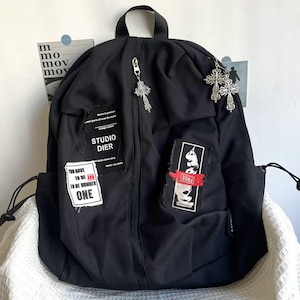Könnte beinhalten: Ein schwarzer Rucksack mit silbernen Reißverschlussanhängern in Kreuzform, einem Tragegriff und zwei Seitentaschen. Die Vorderseite zeigt drei rechteckige Aufnäher mit Texten wie "STUDIO DIER", "YOU HAVE TO BE AND TO BE NUMBER ONE" und "FREE" sowie grafischen Elementen.
