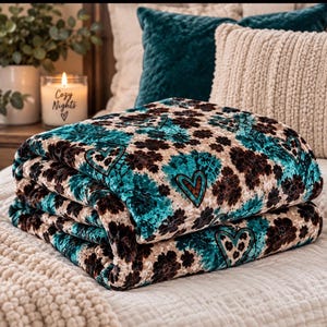 May include: A folded plush blanket featuring a repeating pattern of dark brown floral shapes and teal heart outlines on a cream-colored background. The fabric has a soft, textured appearance with a consistent print throughout the folded layers.