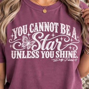 Könnte beinhalten: Ein pflaumenfarbenes T-Shirt mit kurzen Ärmeln und weißem Aufdruck, der YOU CANNOT BE A STAR UNLESS YOU SHINE lautet. Das Design enthält eine Schmetterlingsgrafik, dekorative Sterne und die Signatur Dolly Parton mit einem Herz.