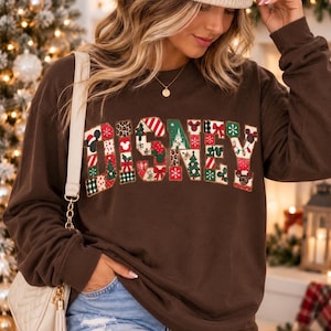 May include: A dark brown long-sleeve sweatshirt featuring the word DISNEY in large block letters. Each letter is filled with a patchwork of holiday-themed patterns including Christmas trees, snowflakes, bows, and Mickey Mouse silhouettes.