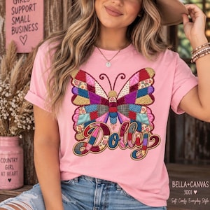 May include: A light pink t-shirt featuring a large, colourful butterfly graphic with a patchwork quilt pattern. The word Dolly is printed below the butterfly in a matching patchwork style. The shirt has short sleeves and a crew neckline.