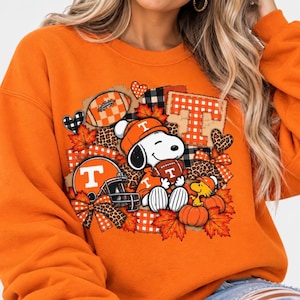 May include: An orange long-sleeve sweatshirt featuring a graphic print of Snoopy wearing an orange beanie and holding a football. The design includes a football helmet with a letter T, pumpkins, autumn leaves, hearts, and plaid patterns.