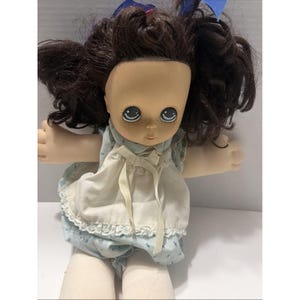 Pode incluir: Uma boneca com cabelo castanho escuro em marias-chiquinhas com fitas azuis, olhos grandes azuis, sardas e pele clara. A boneca veste um vestido azul claro com um avental branco, acabamento em renda e um pequeno laço branco no pescoço.