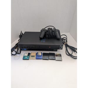 Op de afbeelding: Een zwarte PlayStation 2 spelcomputer met een bijpassende zwarte bedrade controller, stroom- en AV-kabels, en zes diverse geheugenkaarten in kleuren zoals blauw, beige en zwart, allemaal voorzien van zichtbare branding en poortconnectoren.