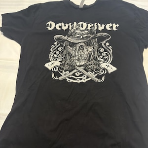May include: A black short-sleeve t-shirt featuring a white graphic print of a skull wearing a cowboy hat. Below the skull are two crossed revolvers and decorative scrollwork. The text DevilDriver is printed in a stylized white font above the skull.