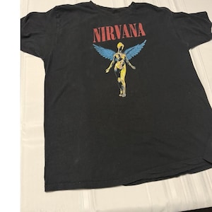 Puede incluir: Una camiseta negra de manga corta con el texto rojo NIRVANA impreso sobre un gráfico de una figura amarilla con grandes alas azules. El diseño está centrado en el pecho de la camiseta, que cuenta con un cuello redondo.