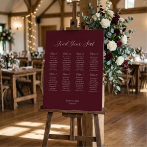 May include: A deep burgundy wedding seating chart sign on a wooden easel, featuring the text "Find Your Seat," guest names organized by tables 1 through 8, and the names "Jessica & Sam" with the date "24.09.2026" in white script and serif fonts.