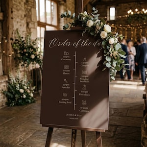 May include: A dark brown wedding order of the day sign on a wooden easel, decorated with white roses and eucalyptus. The sign lists a timeline from 1:00pm Ceremony to 11:30pm Carriages, featuring the names Jessica & Sam and the date 24.09.2026.
