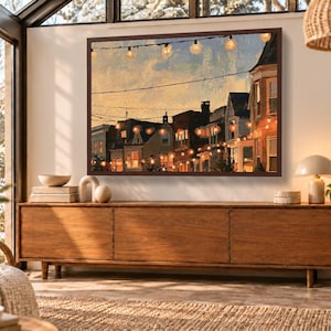 May include: A rectangular framed wall art print depicting a street scene at dusk with glowing string lights hanging between buildings. The painting features warm orange and yellow hues against a twilight sky, showcasing architectural details.