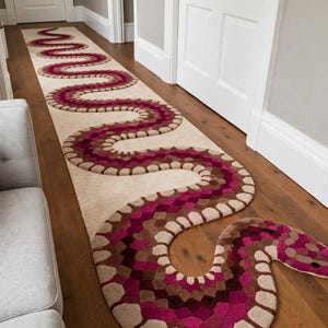 May include: A long, rectangular runner rug featuring a large, winding snake design that spans the entire length. The snake is patterned with shades of magenta, deep pink, and brown, set against a light cream background with a visible forked tongue.