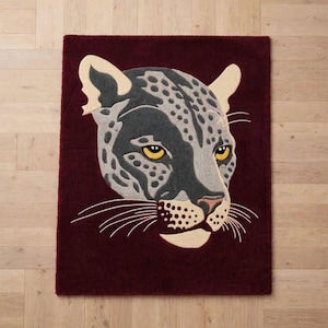 May include: A rectangular rug in deep burgundy featuring a large, centred tufted design of a leopard head. The leopard is rendered in shades of charcoal grey, light grey, and cream, with piercing yellow eyes and long, thin white whiskers.