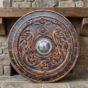 May include: A circular wooden shield featuring intricate carvings of two intertwined serpents and a central hammer symbol. The shield has a polished silver metal boss in the centre and a dark outer border engraved with runic characters.