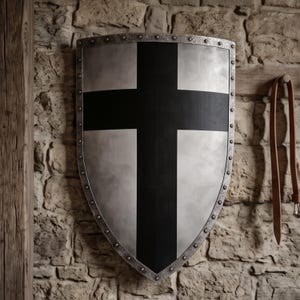 Może przedstawiać: Tarcza typu heraldycznego o metalicznym srebrnym wykończeniu z wyraźnym czarnym krzyżem namalowanym pionowo i poziomo na środku. Tarcza jest otoczona teksturowaną, ciemną metalową krawędzią zabezpieczoną równomiernie rozmieszczonymi nitami.