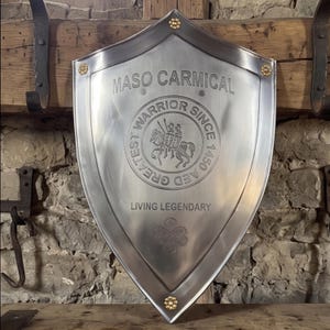 May include: A polished silver-toned metal shield featuring the engraved text MASO CARMICAL, GREATEST WARRIOR SINCE 1450 AED, and LIVING LEGENDARY. The centre displays a circular emblem of a mounted knight, with gold-toned floral rivets at the corners.