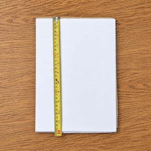 Op de afbeelding: Een rechthoekig wit vel papier dat verticaal is geplaatst. Een geel meetlint is langs de linkerrand geplaatst en toont een lengte van ongeveer 30 cm. Het papier heeft een glad, schoon oppervlak en overal scherpe, rechte randen.