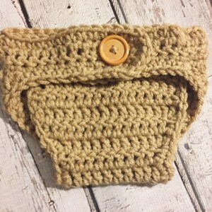 Puede incluir: Una cubierta de pa&ntilde;al de ganchillo de color marr&oacute;n claro con un bot&oacute;n de madera. La cubierta de pa&ntilde;al est&aacute; hecha con un patr&oacute;n de punto simple.