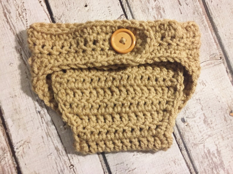 Puede incluir: Una cubierta de pa&ntilde;al de ganchillo de color marr&oacute;n claro con un bot&oacute;n de madera. La cubierta de pa&ntilde;al est&aacute; hecha con un patr&oacute;n de punto simple.