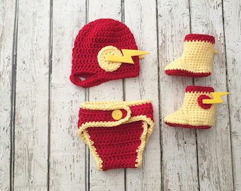 May include: A red and yellow crocheted baby outfit set, including a hat, diaper cover, and two booties. The hat and booties feature a yellow lightning bolt design. The diaper cover has a yellow button closure.