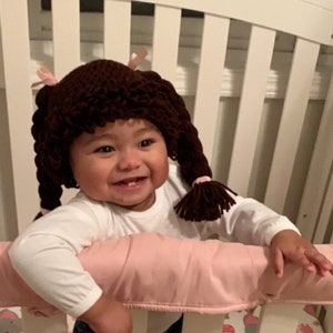 May include: A young child wearing a brown crocheted doll hat with pink bows and brown yarn braids. The child is smiling and looking to the right. The child is standing in a crib with a pink bumper.