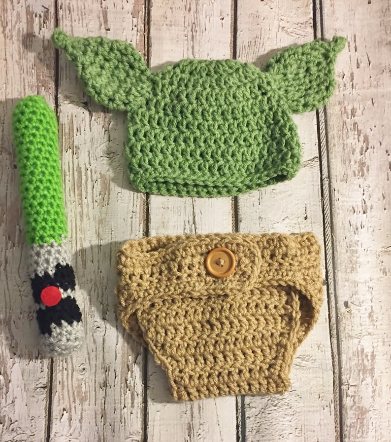 Puede incluir: Un gorro de Yoda de crochet verde con orejas, un cubrepa&ntilde;al de crochet marr&oacute;n con un bot&oacute;n de madera y un sable de luz de crochet verde.