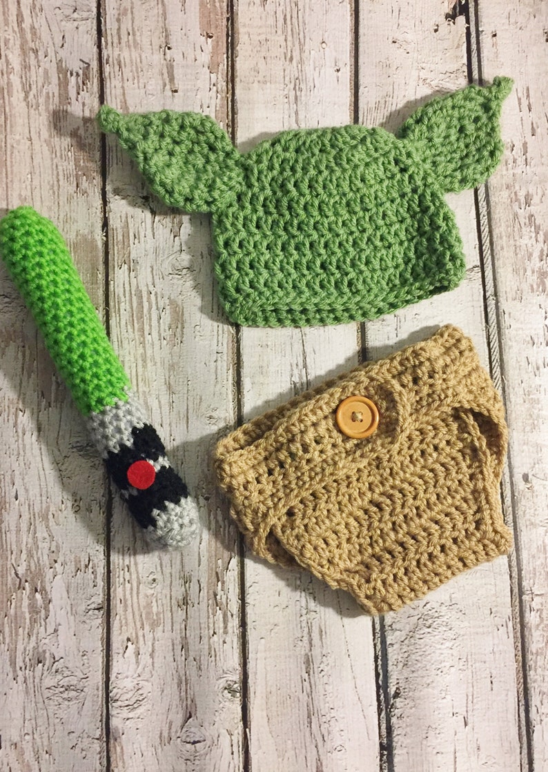 Puede incluir: Un gorro de Yoda de crochet verde con orejas grandes, un pa&ntilde;al de crochet marr&oacute;n con un bot&oacute;n y un sable de luz de crochet verde y gris.