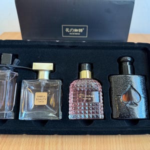 Könnte beinhalten: Ein Set aus vier Parfümflaschen in einem schwarzen Tablett. Die erste ist eine hohe, klare Flasche mit schwarzem Verschluss und Schleife. Die zweite ist eine quadratische, klare Flasche mit goldenem Verschluss und der Aufschrift GABELIL BEAUTY. Die dritte ist eine rosa strukturierte Flasche. Die vierte ist eine schwarze Flasche mit einem Pik-Symbol.