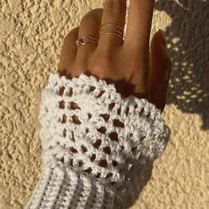Peut inclure: Un gant sans doigts en crochet blanc doté d'un poignet côtelé et d'un motif en dentelle ajourée. L'accessoire est porté au poignet et à la main, complété par trois fines bagues dorées sur les doigts, mettant en valeur un design textile détaillé.