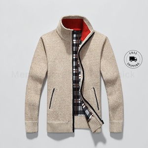 May include: A beige knit zip-up jacket featuring a high collar with a red interior lining. The jacket has long sleeves, ribbed cuffs, and two side pockets with black trim. The interior is lined with a black, white, and red plaid fabric pattern.