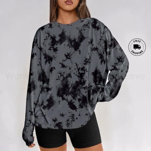 Op de afbeelding: Een sweatshirt met lange mouwen en een ronde hals, voorzien van een grijze basis met een zwart tie-dye spetterpatroon. Het kledingstuk heeft een relaxte pasvorm met geribbelde boorden en een geribbelde halslijn met een abstract patroon.