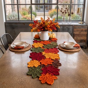 May include: A table runner composed of interconnected crocheted maple leaves in shades of red, orange, yellow, brown, and green. The runner is centered on a table with two place settings, each featuring a woven placemat, a white bowl, and a napkin.