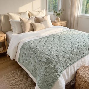 Op de afbeelding: Een saliegroene gebreide plaid met een mandvlechtstructuur gedrapeerd over een wit bedsprei. Het bed is voorzien van meerdere kussens in crème- en beigetinten, gerangschikt tegen een neutraal gestoffeerd hoofdeinde in een lichte slaapkamer.
