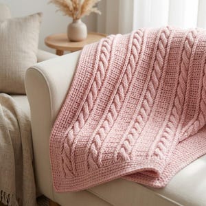 May include: A soft, light pink knitted throw blanket featuring a prominent vertical cable knit pattern alternating with sections of simple garter stitch. The textile has a thick, textured appearance with a consistent pastel color throughout the fabric.