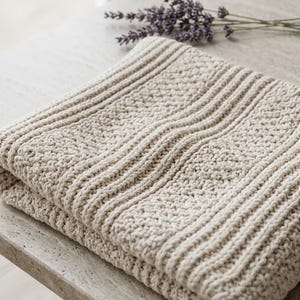 May include: A folded beige knit blanket featuring a textured pattern of alternating vertical ribbed stripes and sections of dense, bumpy stitch work. The fabric has a consistent light tan color throughout, showcasing a thick, soft yarn construction.