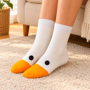 Könnte beinhalten: Ein Paar weiße Crew-Socken mit einem Entengesicht-Motiv. Die Socken haben einen einfarbig weißen Schaft, ein kleines schwarzes kreisförmiges Auge auf jeder Seite und eine leuchtend orangefarbene Zehenpartie, die an einen Entenschnabel erinnert.