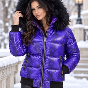 Könnte beinhalten: Eine leuchtend violette Steppjacke mit schwarzem Schlangenledermuster, durchgehendem Frontreißverschluss und schwarzen Rippbündchen an Ärmeln und Saum. Die Jacke verfügt über eine große Kapuze, die mit dickem, schwarzem Kunstfell besetzt ist.