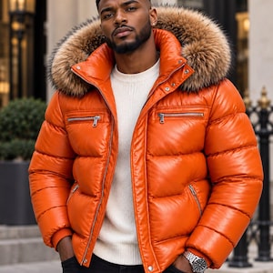 Puede incluir: Una chaqueta acolchada de color naranja brillante con cuello alto, capucha extraíble con ribete de piel sintética marrón, cremalleras metálicas plateadas en el pecho y bolsillos laterales, y puños acanalados, sobre un suéter de punto blanco.