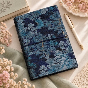 May include: A rectangular notebook featuring a dark blue cover with a textured, intricate pattern of trees and foliage in shades of light blue, teal, and gold. A thin, dark blue elastic band wraps horizontally around the centre of the notebook.