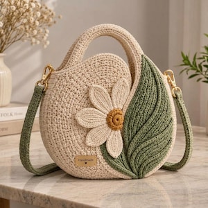 Könnte beinhalten: Eine runde, cremefarbene Häkeltasche mit einer großen, weißen Gänseblümchen-Applikation mit gelber Mitte und einem markanten grünen Blattdesign an der Seite. Die Tasche hat einen passenden grünen Häkel-Schulterriemen mit goldfarbenen Metallbeschlägen.