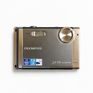 May include: A metallic gray Olympus digital camera featuring a 7.6 cm LCD screen, a 7.1 megapixel sensor, and an all-weather design. The front displays the Olympus brand name, a flash unit, a circular lens, and text indicating high ISO capabilities.