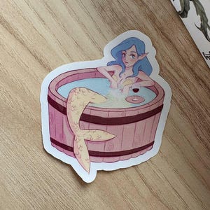 Può includere: Un adesivo fustellato con l'illustrazione di una sirena dai capelli blu e le orecchie a punta che si rilassa in un barile di legno rotondo pieno d'acqua. La sirena ha una coda chiara e tiene in mano un piccolo bicchiere di vino rosso.