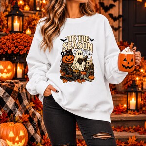 May include: A white long-sleeve sweatshirt featuring a graphic print with the text 'TIS THE SEASON' above a scene of a ghost reading a book, a black cat on a pumpkin, a witch hat, a cauldron, a skull, and various autumn-themed elements.