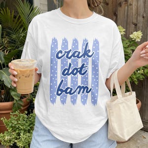 May include: A white short-sleeve t-shirt featuring a graphic of vertical light blue brushstrokes with white polka dots and the dark blue text "crak dot bam" centred on the front. The shirt is paired with light blue and white striped shorts.